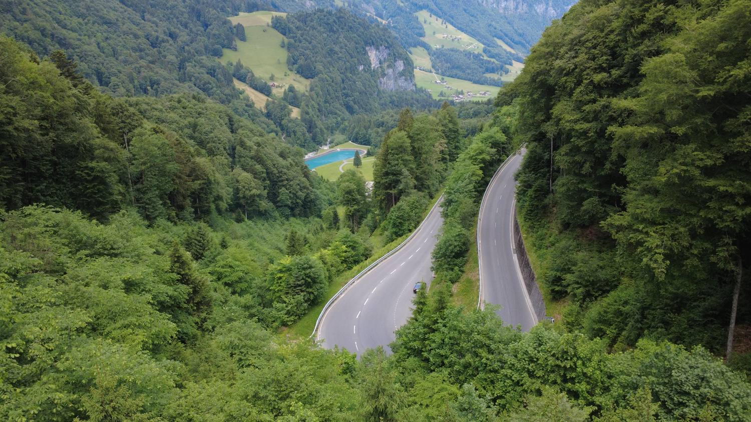 The hairpin bends of the Brünig Pass road climbing through the forest, with a reservoir and hamlets in the valley below
