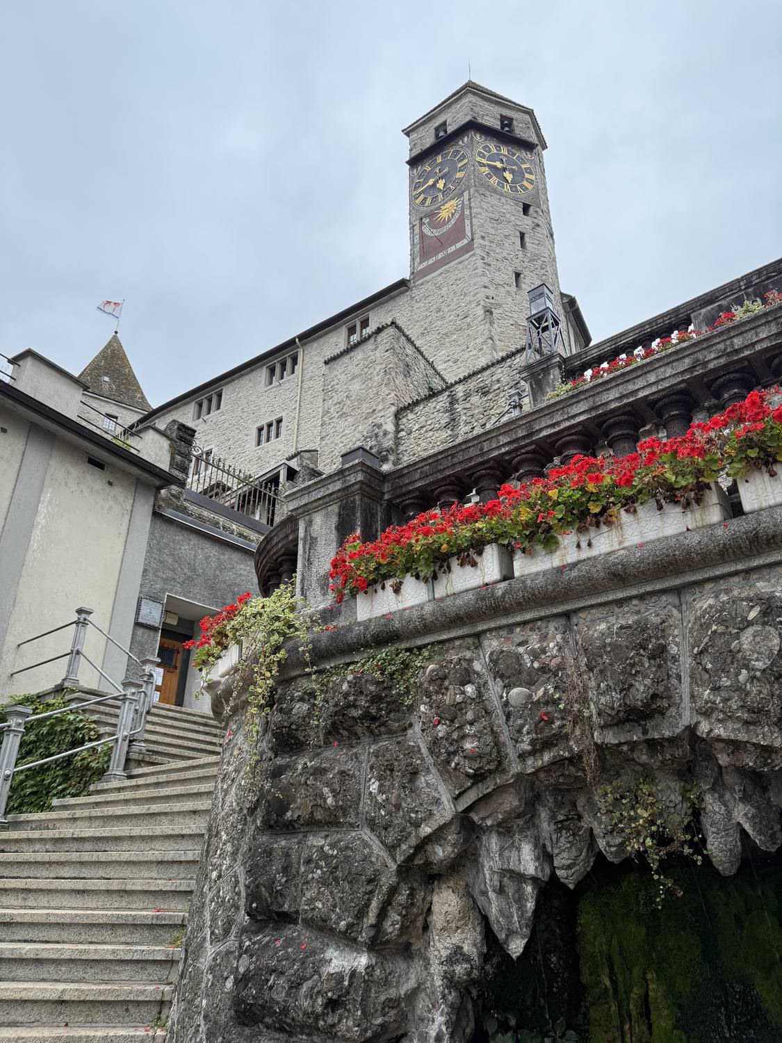 The clock tower of Rapperswil Castle above a stairway lined with red geraniums